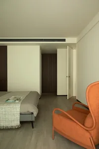 A minimalist bedroom with light wooden flooring, a grey bed, and an orange chair.