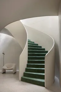 Spiraling staircase with green carpeted steps and white concrete walls.