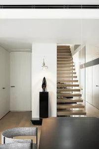 A minimalist interior featuring a wooden staircase with floating steps and an abstract black sculpture on a pedestal.