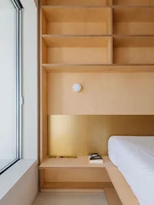 A minimalist bedroom featuring a wooden storage unit within the headboard area, next to a large window.