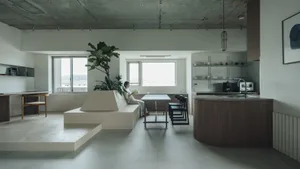 A modern kitchen with concrete and wooden finishes, featuring a plant on a elevated platform and a dining area with a woman r