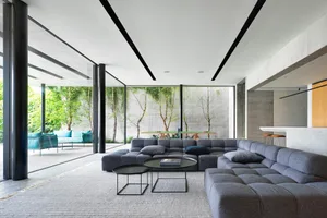 A modern minimalist living room featuring sleek grey furniture, glass walls, and concrete elements, with a view of greenery o