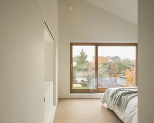 Minimalist bedroom with a large window overlooking a garden, featuring a bed with a white blanket and a wooden floor.