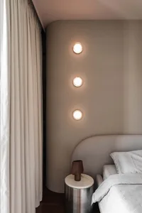 A minimalist bedroom with a textured grey headboard, white bedding, and a sleek, modern bedside table with a vase.
