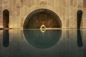 A man sitting on the edge of a swimming pool with an arched wall behind him made of stone.
