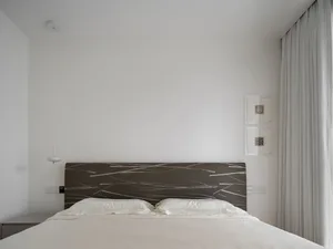 A minimalist bedroom featuring a bed with a patterned headboard and white bedding, alongside a white pendant light and a wind