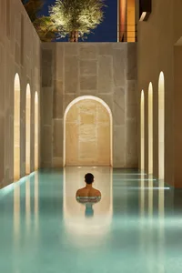 A serene swimming pool with a modern design and concrete walls, featuring a lone figure sitting in the pool.