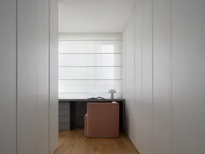 A minimalist home office studio featuring a pink chair, a desk with a lamp, and a large window with white blinds.