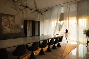 A modern living and dining room with a long marble table, glass chandelier, and sheer curtains.
