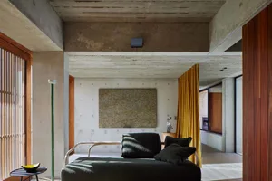 A modern industrial living room featuring bare concrete walls, a dark green lounge chair, and a yellow curtain.