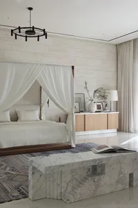A modern minimalist bedroom featuring a canopy bed draped with white fabric, a marble bench, wooden drawers, and a textured r