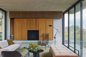 A modern living room featuring a wooden fireplace, concrete walls, and large windows overlooking the outdoors.