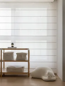 A minimalist interior featuring fabric blinds, storage bins, and a plush cloud-shaped cushion on a wooden floor.