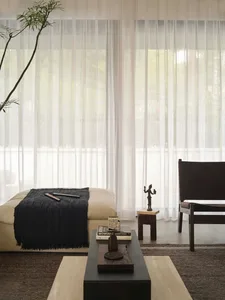 A minimalist living room with a beige sofa, black curtains, a black ottoman, and a black and wood coffee table.