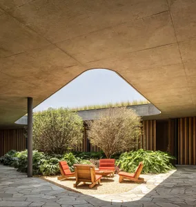 A modern indoor lounge area featuring wooden chairs on a gravel surface surrounded by greenery, with an arched roof and natur