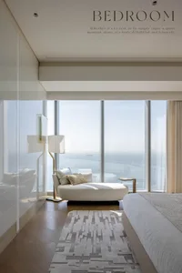 A serene minimalist bedroom featuring white accents, a large window with a sea view, and a comfortable seating area on a patt