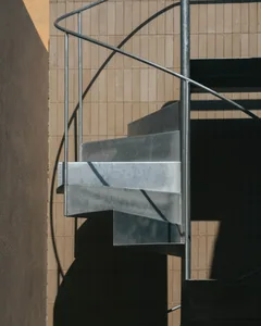 A rugged, metallic spiral staircase contrasts with a smooth, concrete building exterior.