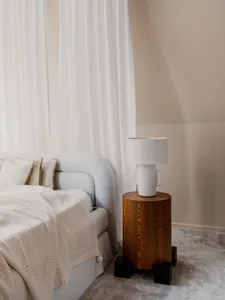 A minimalist bedroom featuring a white bed with pillows, a beige blanket, and a wooden side table with a ceramic lamp. White