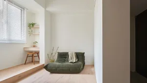 A modern minimalist living room with a green plush sofa, a wooden stool under a white window with blinds.