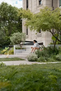 A woman sits on concrete steps surrounded by grass and trees in an urban garden setting.