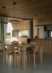 A modern dining room with a wooden table and chairs, featuring large windows and a kitchen counter.