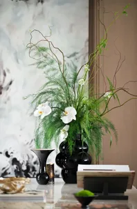 A black vase with white orchids, greenery, and spiral branches on a reflective surface.