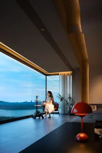 A minimalist living room featuring wooden accents and glass walls, with a woman sitting on a chair near a large window.