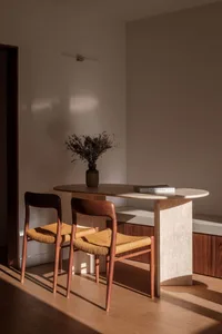 A minimalist dining room with a wooden table and chairs, featuring a vase of dried flowers and a book.