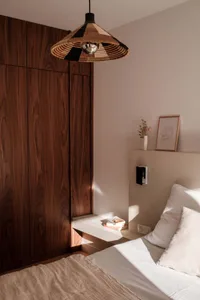 A minimalist bedroom featuring a wooden wardrobe, a bed with a plain white mattress and pillows, and a bedside table with a v
