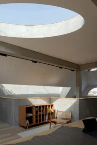 Modern minimalist bedroom featuring a concrete ceiling with a large circular cutout, natural light, and simple furniture.