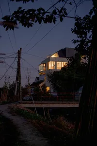 A modern residential house with a steel frame illuminated at night, viewed from a road with a bridge over a stream.
