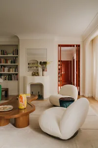 A cozy living room with white upholstered chairs, wooden coffee table, and bookshelves filled with books.