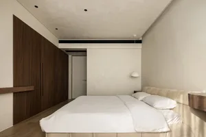 A minimalist bedroom featuring a white bed with a beige suede headboard, a dark wood closet, and a white ceiling light fixtur