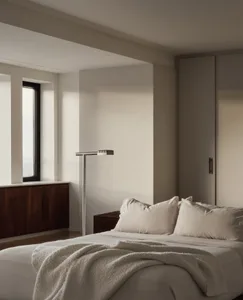 A minimalist bedroom with neutral-colored walls, bedding, and pillows, featuring a simple floor lamp and a window with a view