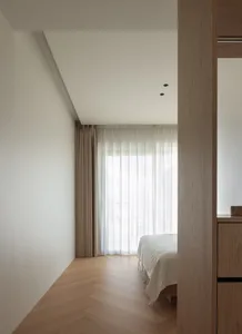 A modern minimalist bedroom with wooden flooring and light-colored curtains.