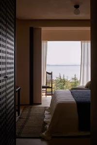 A minimalist bedroom with a bed, chair, and view of the water and trees.