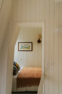 A minimalist bedroom with a bed covered in white sheets and a rust-colored blanket, featuring wooden beadboard walls and a fr