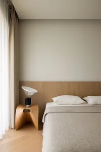 A minimalist bedroom featuring a wooden bedside lamp, a fabric bedspread, and a minimalistic headboard made of wood.