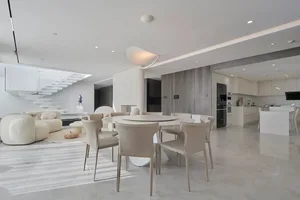 A modern living, dining, and kitchen area with white marble floors, a white round dining table surrounded by beige chairs, an