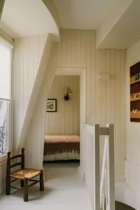 A minimalist bedroom with white wooden walls and a bed covered in a brown blanket, featuring a small wooden chair and a frame