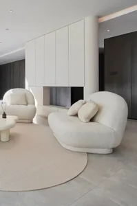 Modern white living room featuring two white velvet armchairs on a round beige rug, minimalist white cabinets, and a fireplac