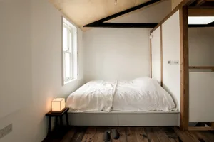 A minimalist white bedroom with a bed on a raised platform, wooden floor, and simple bedside lamp.