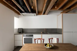 A minimalist residential kitchen featuring a concrete countertop and floor, exposed wooden beams, and modern appliances.