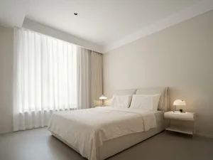 Modern minimalist bedroom with light-colored bedding and curtains on a large window.