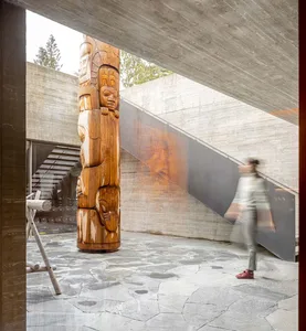 A modern indoor space featuring concrete decoration with a wooden totem pole, stone floor tiles, and a person walking on the 