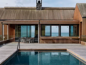 A modern minimalist house with a wooden exterior, large windows overlooking the ocean, and a swimming pool.