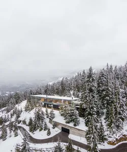 A modern, glass-fronted residence built on a snowy, forested hillside.
