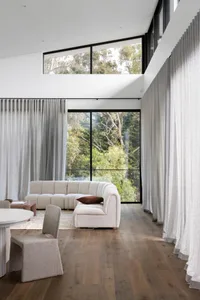 Modern minimalist living room with white fabric sectional sofa, wood flooring, and large windows overlooking greenery.