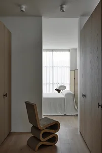 A minimalist bedroom featuring a wooden lamp, white bedding, and a curved chair.