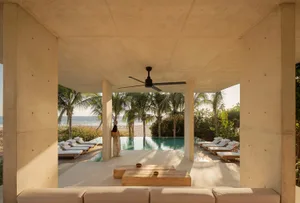 A modern minimalist residential patio with concrete pillars, lounge chairs, and an infinity pool overlooking the sea and palm